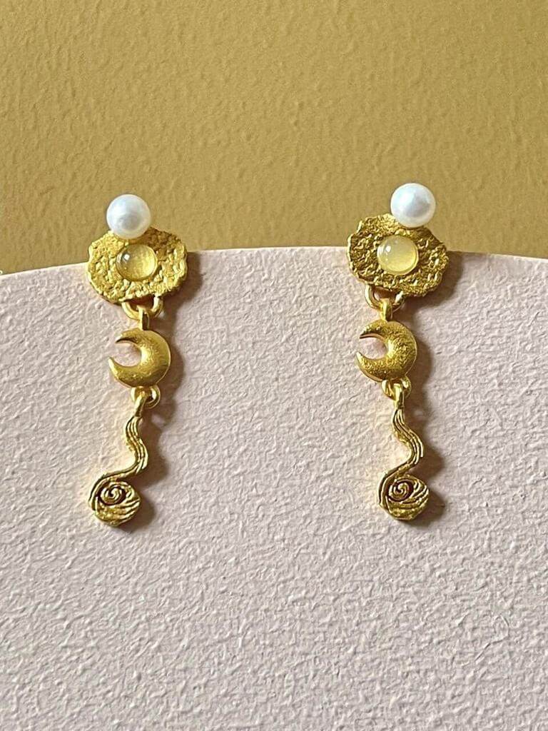 Pallantis Måne River Øreringe er guldfarvede hængeøreringe med teksturerede cirkulære toppe, hver prydet med en hvid perle, rund sten, måne vedhæng og spiralformet hvirvelvedhæng hængende nedenunder.